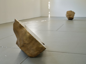 Two boulder-like sculptures, one with a flat surface visible in profile, sit far from each other on the floor of a large gallery with white walls.